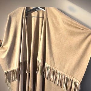 May include: A tan open-front poncho with long sleeves and a fringed hem. The poncho is made of a soft, suede-like material and hangs on a hanger. The fringe is a lighter shade of tan.