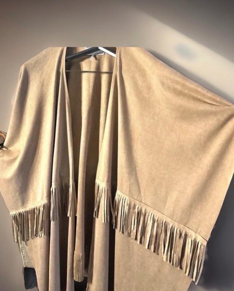 May include: A tan open-front poncho with long sleeves and a fringed hem. The poncho is made of a soft, suede-like material and hangs on a hanger. The fringe is a lighter shade of tan.