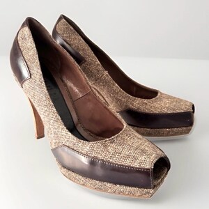 May include: A pair of peep-toe high heels featuring a textured brown tweed fabric upper combined with smooth dark brown leather trim along the base and heel counter, set on a high stiletto heel with a platform sole.
