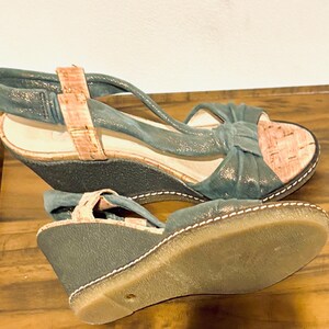 May include: A pair of teal wedge sandals with cork accents. The sandals feature a bow detail on the front and a strap that wraps around the ankle. The soles are a light brown color with white stitching.
