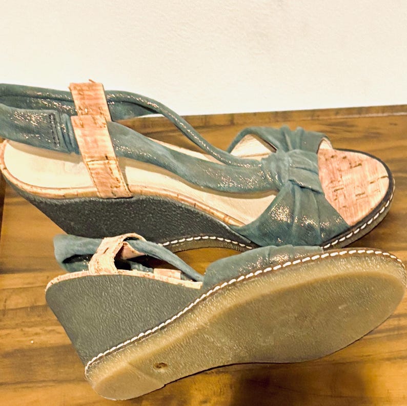 May include: A pair of teal wedge sandals with cork accents. The sandals feature a bow detail on the front and a strap that wraps around the ankle. The soles are a light brown color with white stitching.