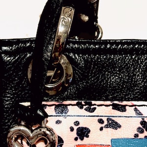 Pode incluir: Bolsa de couro preta com ferragens prateadas, um berloque em forma de cora&ccedil;&atilde;o e uma al&ccedil;a. A bolsa apresenta uma se&ccedil;&atilde;o estampada em rosa e branco com pegadas pretas e as letras "ST" em vermelho e azul.