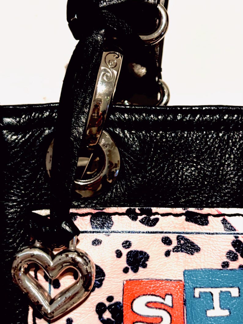 Pode incluir: Bolsa de couro preta com ferragens prateadas, um berloque em forma de cora&ccedil;&atilde;o e uma al&ccedil;a. A bolsa apresenta uma se&ccedil;&atilde;o estampada em rosa e branco com pegadas pretas e as letras "ST" em vermelho e azul.