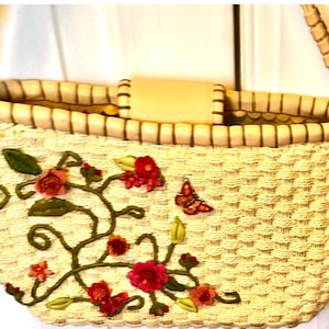 Puede incluir: Un bolso tote de paja tejido con un asa de cuero marr&oacute;n y un dise&ntilde;o floral bordado con flores rosas, rojas y verdes y una mariposa.