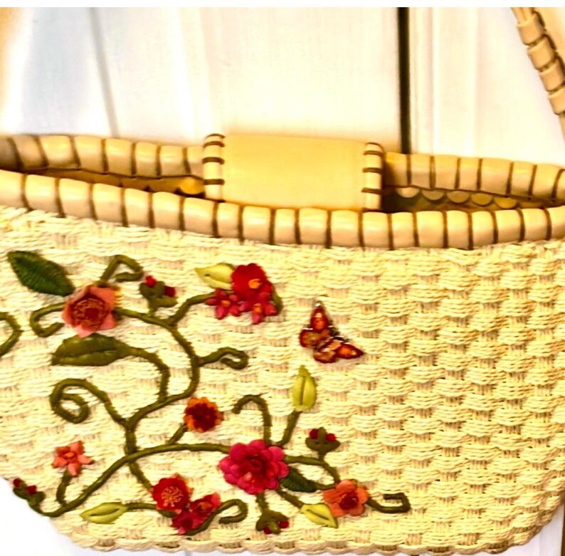 Puede incluir: Un bolso tote de paja tejido con un asa de cuero marr&oacute;n y un dise&ntilde;o floral bordado con flores rosas, rojas y verdes y una mariposa.