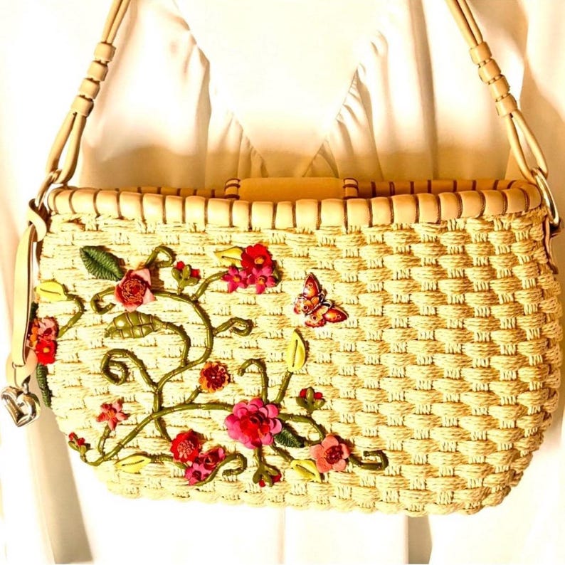 Puede incluir: Un bolso de paja tejido con una correa de cuero marr&oacute;n claro y bordado floral. El bordado presenta flores y hojas rosas, rojas y verdes, as&iacute; como una mariposa.