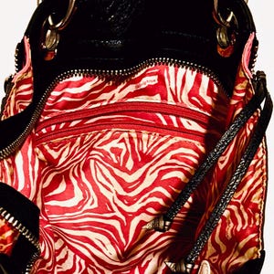 Pode incluir: Bolsa de couro preta com detalhes prateados e forro com estampa de zebra vermelha e branca. A bolsa tem v&aacute;rios compartimentos com z&iacute;per e uma al&ccedil;a de ombro. O interior tem a palavra "Brighton" impressa.