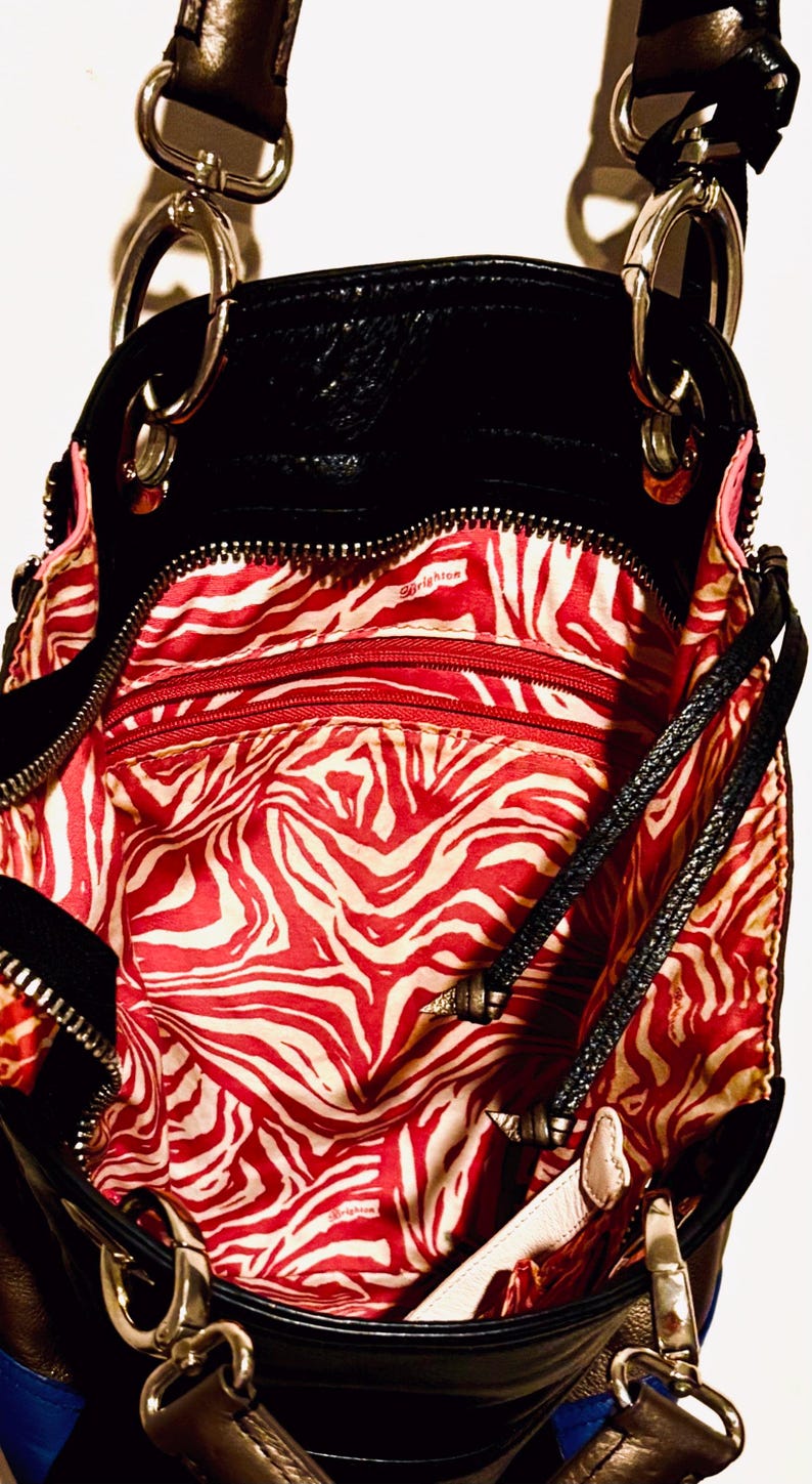 Pode incluir: Bolsa de couro preta com detalhes prateados e forro com estampa de zebra vermelha e branca. A bolsa tem v&aacute;rios compartimentos com z&iacute;per e uma al&ccedil;a de ombro. O interior tem a palavra "Brighton" impressa.
