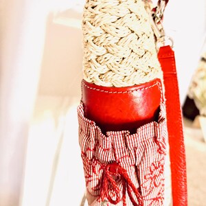 May include: A red and cream-coloured water bottle holder with a woven design. The holder has a red leather top and bottom, with a red and white striped fabric pocket. A red strap is attached to the holder.
