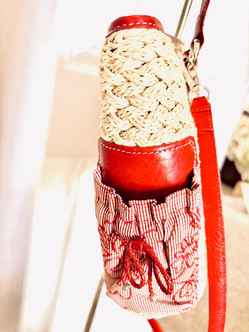 May include: A red and cream-coloured water bottle holder with a woven design. The holder has a red leather top and bottom, with a red and white striped fabric pocket. A red strap is attached to the holder.
