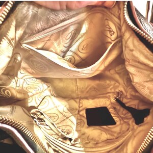 May include: Interior view of a brown handbag with a gold-colored lining. The lining features a repeating pattern of a stylized letter "B" and swirls. The bag has multiple pockets and a zipper closure. A small black tag and a key fob are visible.