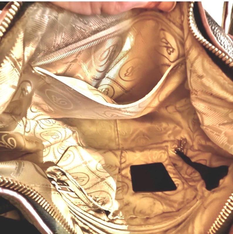 May include: Interior view of a brown handbag with a gold-colored lining. The lining features a repeating pattern of a stylized letter "B" and swirls. The bag has multiple pockets and a zipper closure. A small black tag and a key fob are visible.