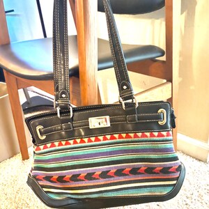 May include: Black handbag with a colorful woven fabric design. The bag features a rectangular shape with a black leather top and handles. The woven fabric has red, green, purple, and blue stripes and geometric patterns. The bag has silver hardware.