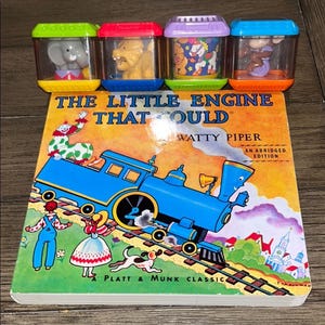 May include: A children's book titled "The Little Engine That Could" by Watty Piper, with a blue train illustration. Above the book are four colorful toy blocks, each containing a different animal figure.
