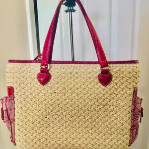 May include: A woven straw tote bag with red detailing. The bag has red handles, trim, and side pockets. Heart-shaped embellishments are on the handle attachments. The bag is suspended from a metal hanger.