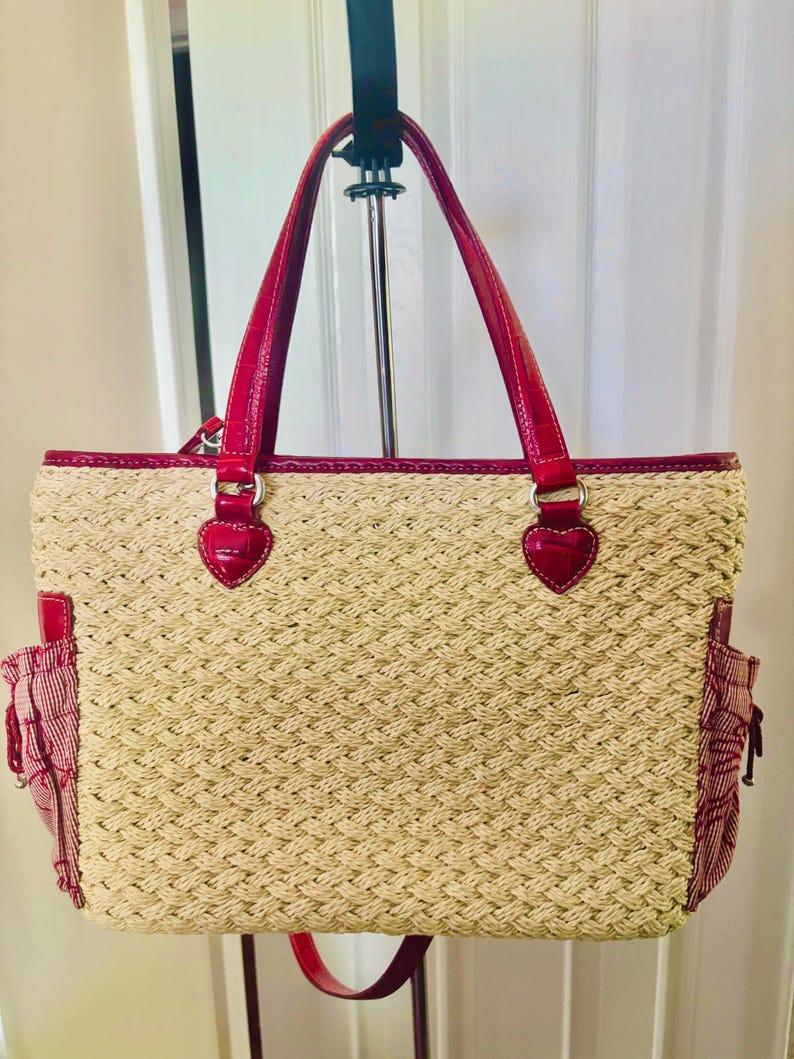 May include: A woven straw tote bag with red detailing. The bag has red handles, trim, and side pockets. Heart-shaped embellishments are on the handle attachments. The bag is suspended from a metal hanger.