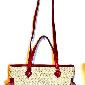 May include: A woven straw tote bag with red leather handles and trim. The bag has a rectangular shape with a textured weave pattern and two small side pockets. The handles are long enough to be worn over the shoulder.
