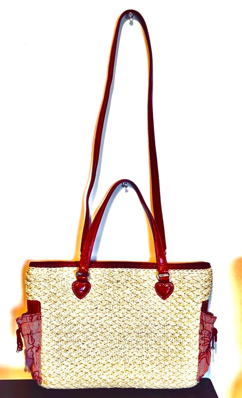 May include: A woven straw tote bag with red leather handles and trim. The bag has a rectangular shape with a textured weave pattern and two small side pockets. The handles are long enough to be worn over the shoulder.