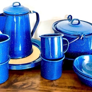 May include: A blue enamelware camping cookware set. The set includes a coffee pot, mugs, plates, a pot with a lid, and bowls. The blue color has white speckles, giving it a vintage look. The set is arranged on a wooden surface.