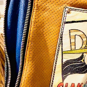 May include: A tan perforated leather jacket featuring a blue inner lining and a silver zipper. A rectangular patch is stitched onto the front, displaying the text GLAM GIRL in red lettering, along with a partial graphic illustration of a figure.