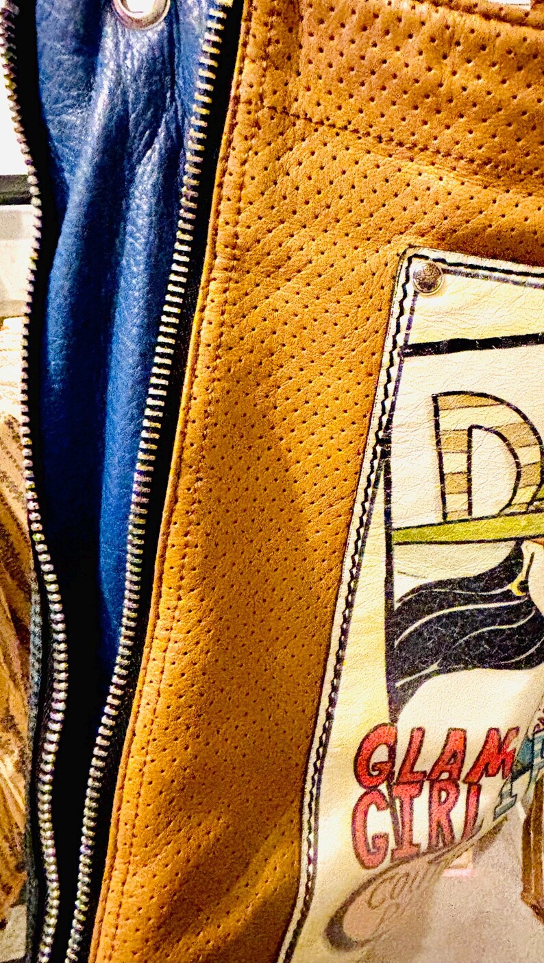 May include: A tan perforated leather jacket featuring a blue inner lining and a silver zipper. A rectangular patch is stitched onto the front, displaying the text GLAM GIRL in red lettering, along with a partial graphic illustration of a figure.