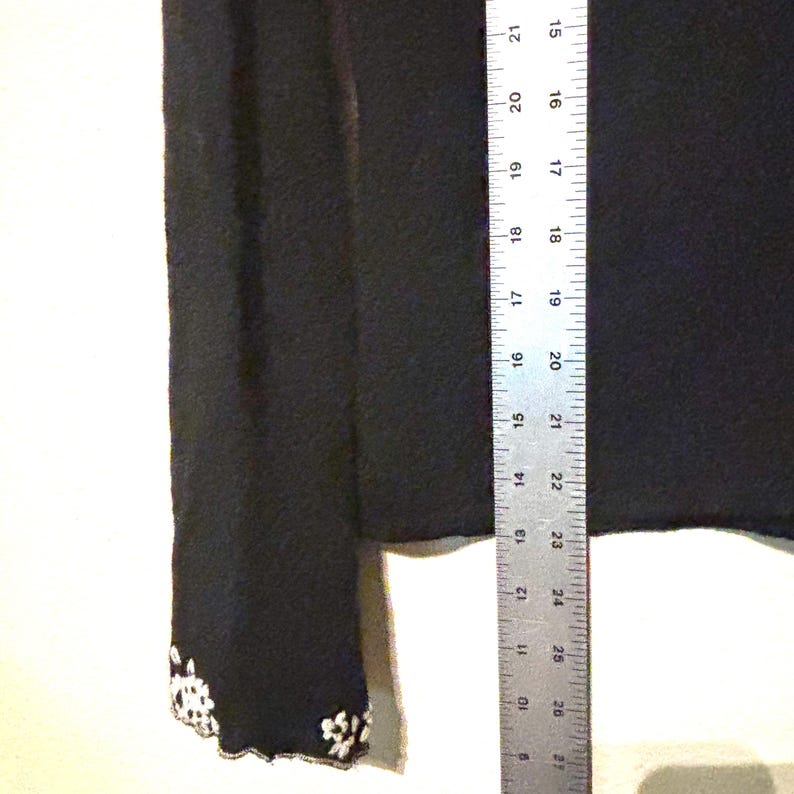 May include: Black long-sleeve garment with white floral embroidery on the cuff. A ruler is placed next to the garment, showing measurements in inches. The garment's fabric appears to be a soft material.