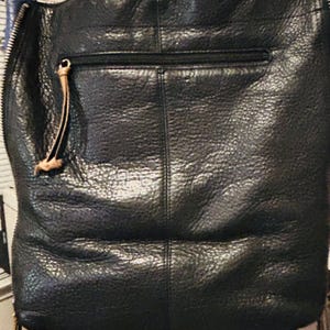 May include: A black textured bag featuring a vertical seam down the center, a horizontal zippered pocket with a tan leather pull tab, and a prominent silver-toned metal zipper running along the left side edge of the bag's exterior surface.