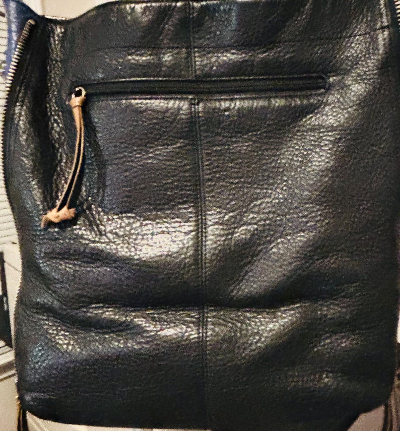 May include: A black textured bag featuring a vertical seam down the center, a horizontal zippered pocket with a tan leather pull tab, and a prominent silver-toned metal zipper running along the left side edge of the bag's exterior surface.