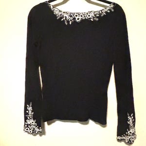 May include: A black long-sleeve sweater with white floral embroidery around the neckline and cuffs. The sweater is hanging on a hanger against a plain background. The embroidery features delicate floral and leaf patterns.