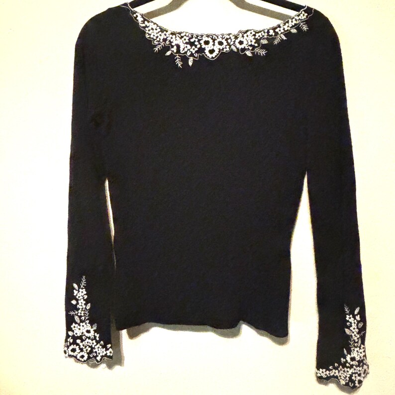May include: A black long-sleeve sweater with white floral embroidery around the neckline and cuffs. The sweater is hanging on a hanger against a plain background. The embroidery features delicate floral and leaf patterns.