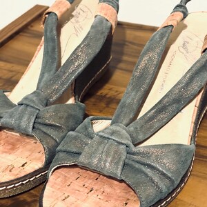 May include: A pair of open-toe wedge sandals. The sandals feature a teal-colored fabric with a bow detail on the toe strap and a cork footbed. The straps are a teal color with a pinkish-tan accent. The soles are dark brown with white stitching.