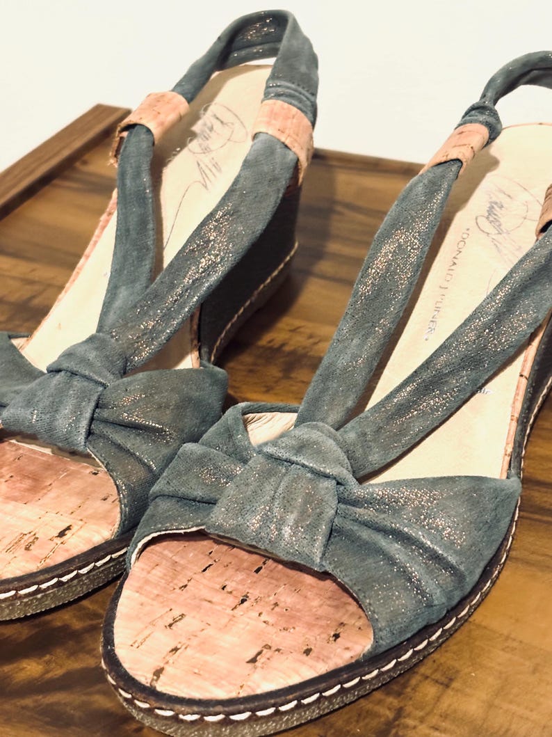 May include: A pair of open-toe wedge sandals. The sandals feature a teal-colored fabric with a bow detail on the toe strap and a cork footbed. The straps are a teal color with a pinkish-tan accent. The soles are dark brown with white stitching.