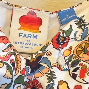May include: A white dress with a colorful floral print. The dress has a V-neck and is made by FARM for Anthropologie. The tag says "Made in Turkey" and "S".