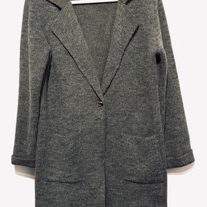 Könnte beinhalten: Ein grauer Strickblazer mit fallendem Revers, langen Ärmeln und zwei Vordertaschen. Der Blazer hat einen einzelnen Knopfverschluss und hängt an einem weißen Kleiderbügel. Der Stoff scheint ein weicher, strukturierter Strick zu sein.