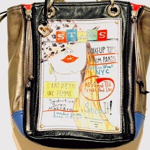 Pode incluir: Bolsa preta, dourada e azul com um gr&aacute;fico do rosto de uma mulher e texto como "STARS", "MAKEUP TIPS FROM PARIS" e "Shop It!". A bolsa tem ferragens prateadas, z&iacute;peres e um pingente em forma de cora&ccedil;&atilde;o.