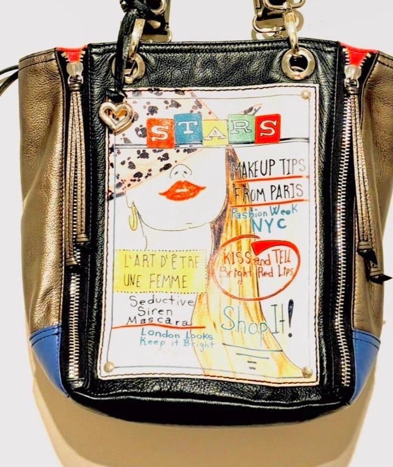 Pode incluir: Bolsa preta, dourada e azul com um gr&aacute;fico do rosto de uma mulher e texto como "STARS", "MAKEUP TIPS FROM PARIS" e "Shop It!". A bolsa tem ferragens prateadas, z&iacute;peres e um pingente em forma de cora&ccedil;&atilde;o.