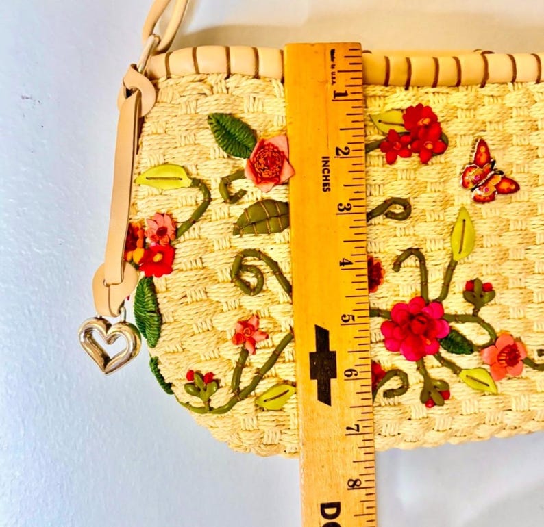 Puede incluir: Un bolso de paja con un dije de coraz&oacute;n y bordado floral. El bolso mide 17,8 cm de largo y presenta flores rosas, rojas y verdes.