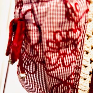 May include: Close-up of a fabric item with a red and white pattern. The fabric has a gingham pattern at the top and a vertical striped pattern below. A red drawstring with a silver bead is visible.