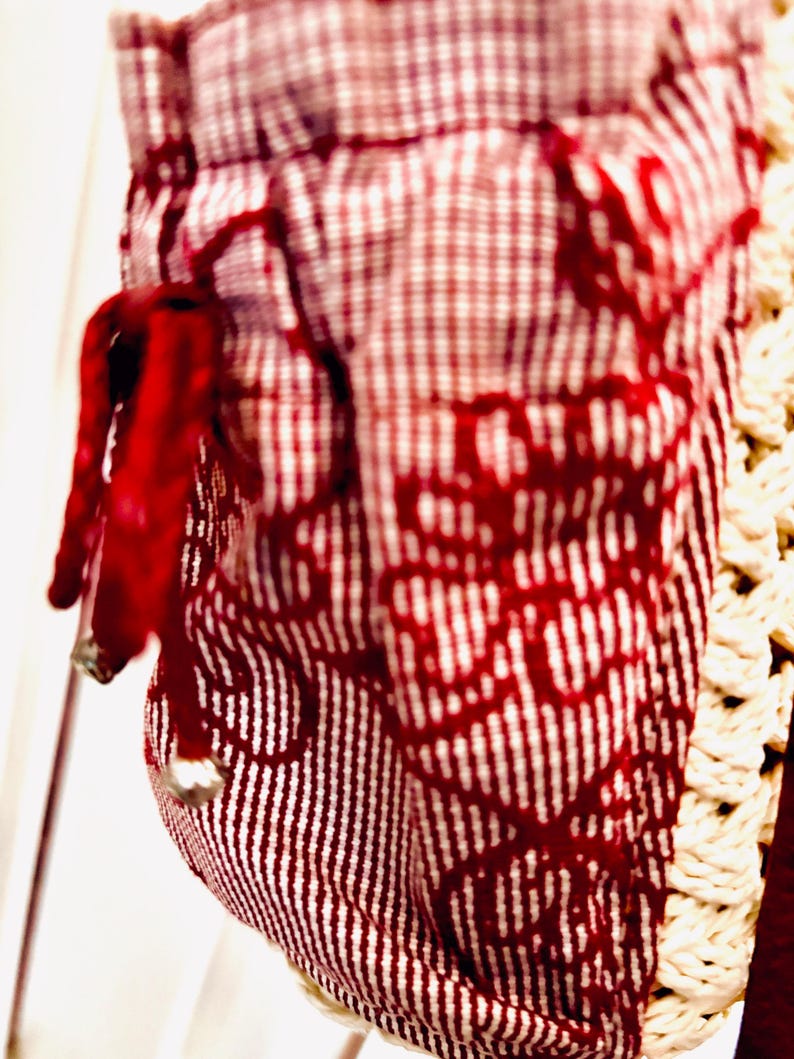 May include: Close-up of a fabric item with a red and white pattern. The fabric has a gingham pattern at the top and a vertical striped pattern below. A red drawstring with a silver bead is visible.