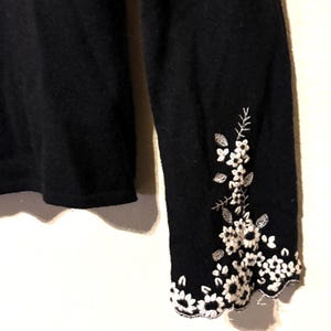 May include: Close-up of a black sweater sleeve with white floral embroidery. The embroidery features detailed flowers and leaves, creating a delicate pattern along the cuff. The sweater's fabric appears soft and textured.
