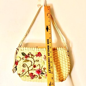 Puede incluir: Un bolso de paja tejido con una correa de cuero blanco y bordado floral. El bordado presenta flores y enredaderas rosas, rojas y verdes con una mariposa.