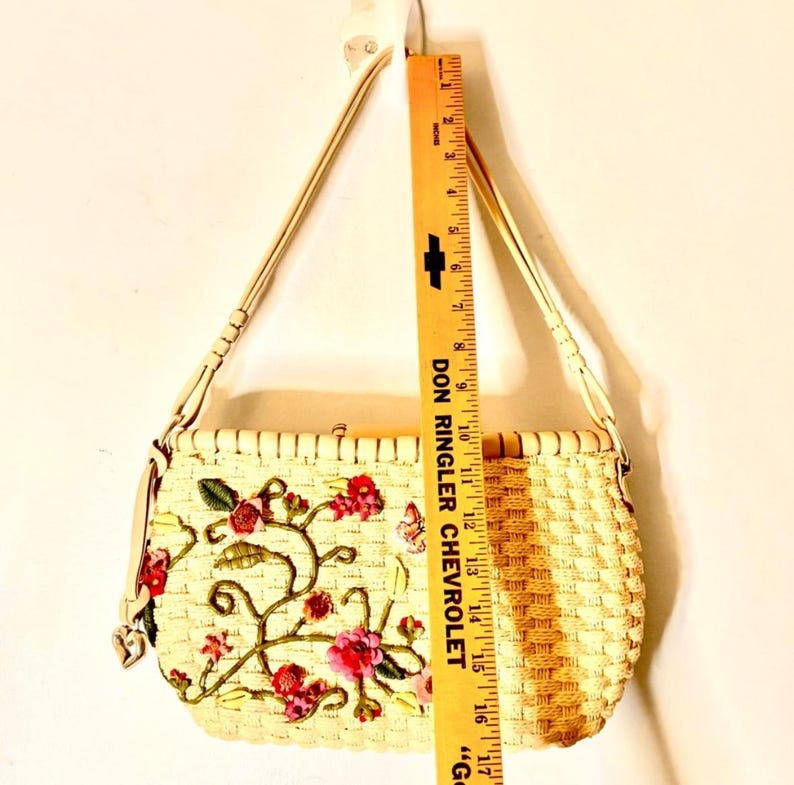 Puede incluir: Un bolso de paja tejido con una correa de cuero blanco y bordado floral. El bordado presenta flores y enredaderas rosas, rojas y verdes con una mariposa.