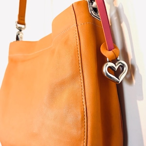 以下が含まれることがあります： シルバーのハート型チャームが付いた、タン色のレザーハンドバッグ。バッグには、シルバーのバックルが付いたショルダーストラップと、ピンクのアクセントストラップが付いています。バッグは滑らかな革でできており、シンプルなデザインです。