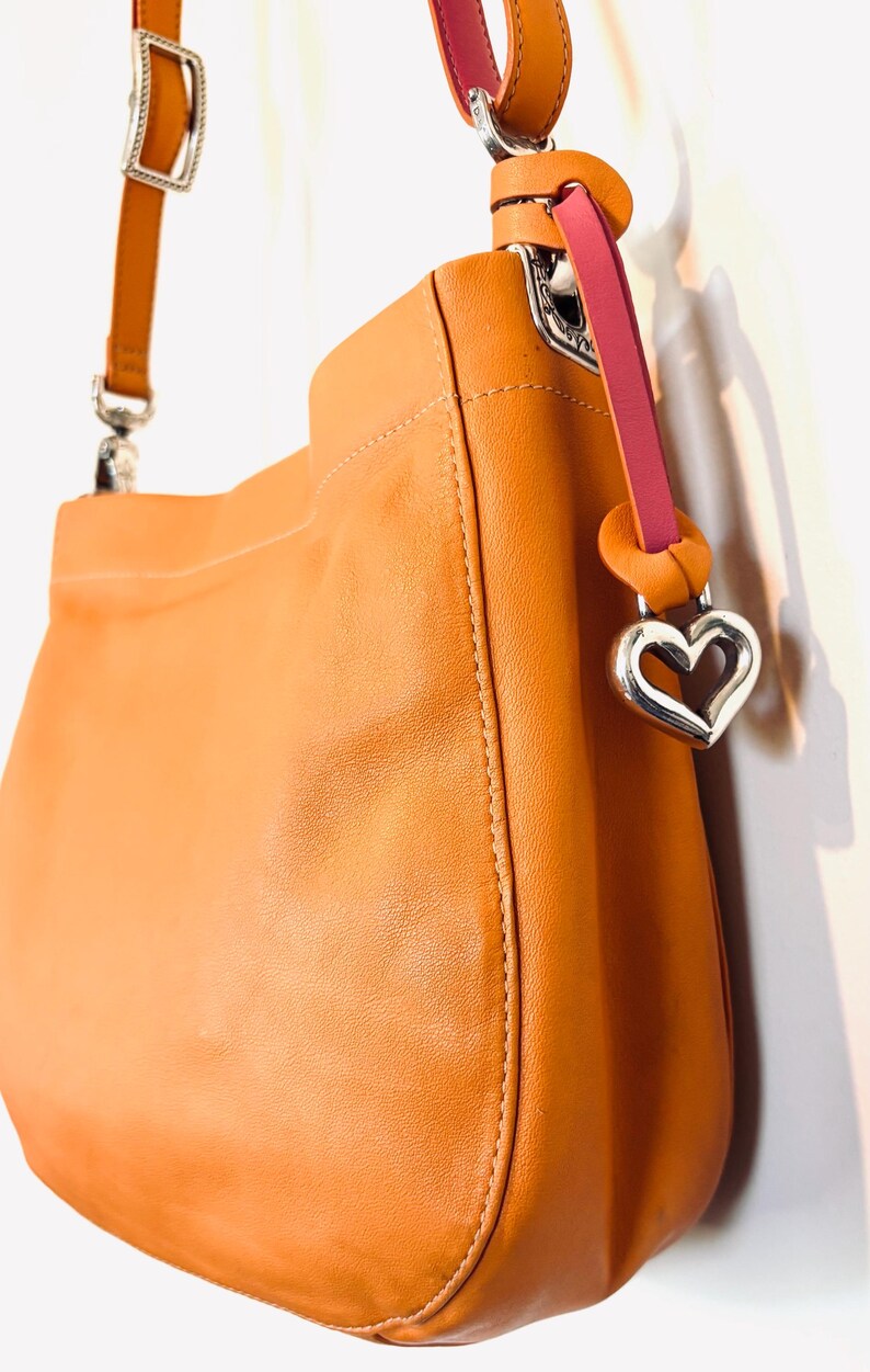 以下が含まれることがあります： シルバーのハート型チャームが付いた、タン色のレザーハンドバッグ。バッグには、シルバーのバックルが付いたショルダーストラップと、ピンクのアクセントストラップが付いています。バッグは滑らかな革でできており、シンプルなデザインです。