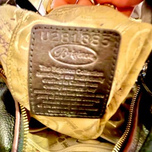 May include: Close-up of a dark brown leather tag inside a handbag. The tag has the number "U281385" embossed at the top, with the brand name "Brighton" below. Text on the tag reads "The Brighton Collection handbags are individually crafted in China from quality leather."