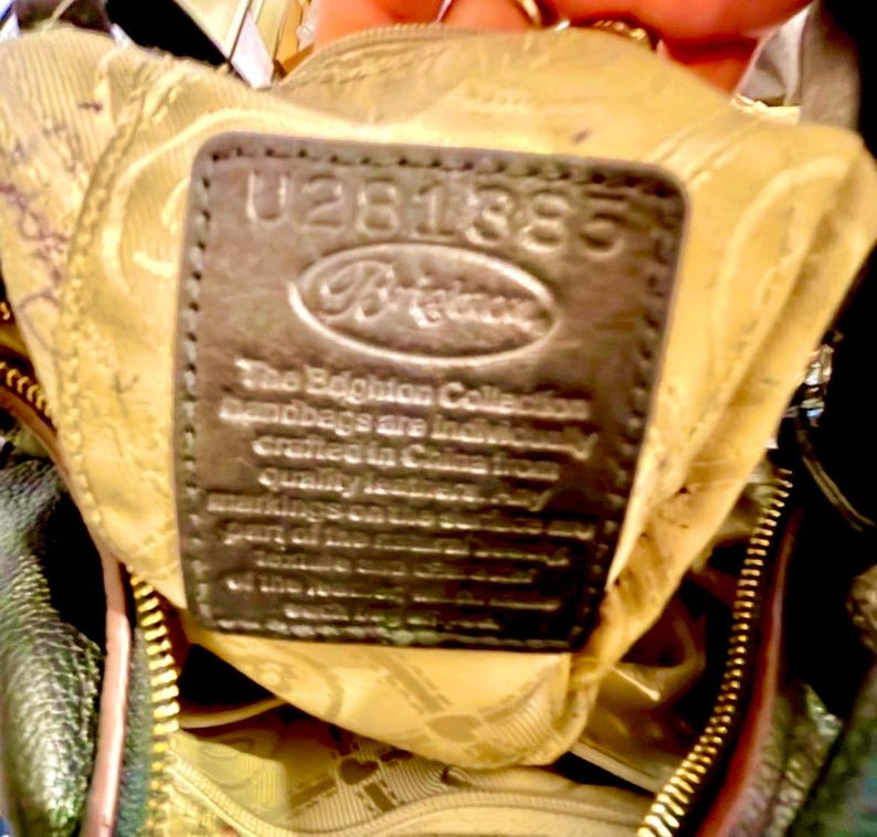 May include: Close-up of a dark brown leather tag inside a handbag. The tag has the number "U281385" embossed at the top, with the brand name "Brighton" below. Text on the tag reads "The Brighton Collection handbags are individually crafted in China from quality leather."