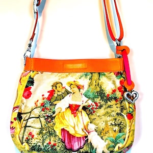 以下が含まれることがあります： 牧歌的なシーンのプリントが施されたカラフルなクロスボディバッグ。バッグは鮮やかなオレンジ色のレザートリムとストラップ、ピンクのアクセント、ハート型のチャームが特徴です。バッグの本体には、ヴィンテージスタイルのイラストが表示されています。
