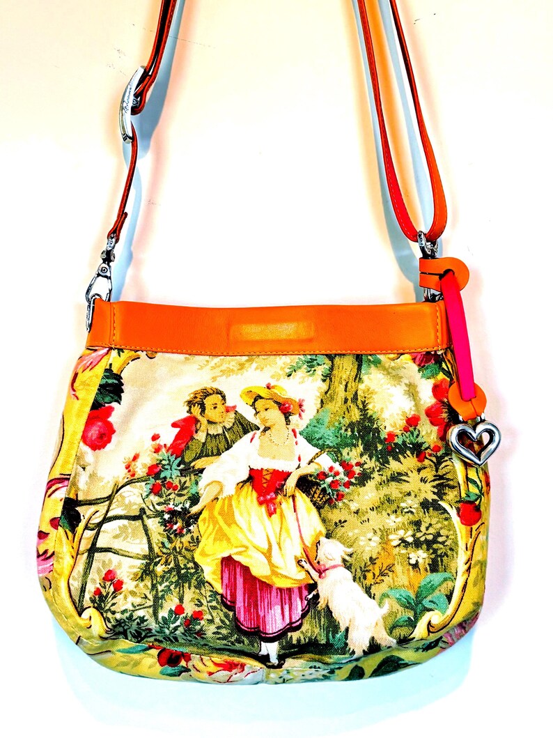 以下が含まれることがあります： 牧歌的なシーンのプリントが施されたカラフルなクロスボディバッグ。バッグは鮮やかなオレンジ色のレザートリムとストラップ、ピンクのアクセント、ハート型のチャームが特徴です。バッグの本体には、ヴィンテージスタイルのイラストが表示されています。