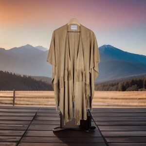 May include: A tan suede poncho with fringe detailing hangs on a wooden hanger. The poncho is displayed against a backdrop of mountains, a wooden deck, and a sunset.