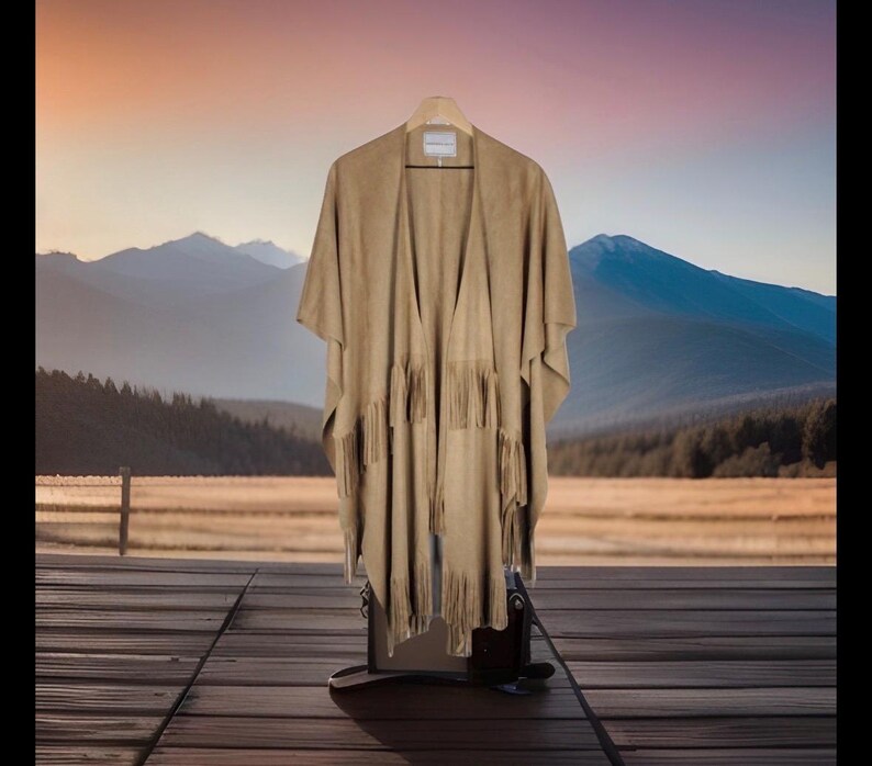 May include: A tan suede poncho with fringe detailing hangs on a wooden hanger. The poncho is displayed against a backdrop of mountains, a wooden deck, and a sunset.